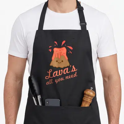 Lavas kawaii apron - Smiling cartoon volcano with coral lava and text Lava's all you need — shown on a black cotton canvas apron against a white background