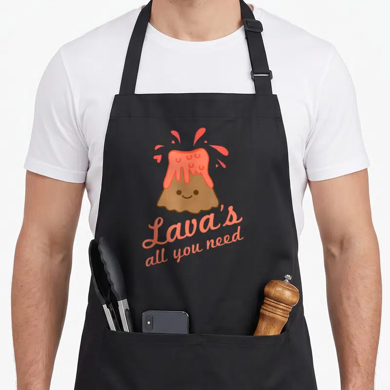 Lavas kawaii apron - Smiling cartoon volcano with coral lava and text Lava's all you need — shown on a black cotton canvas apron against a white background