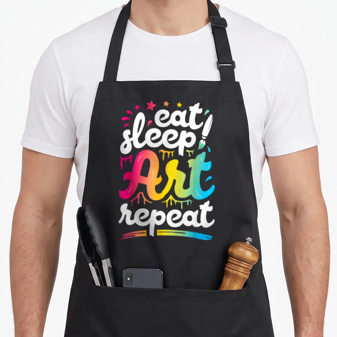 Eat creative apron - Stacked text eat sleep Art repeat with rainbow gradient, paint splatters, stars, and paintbrush — shown on a black cotton canvas apron against a white background