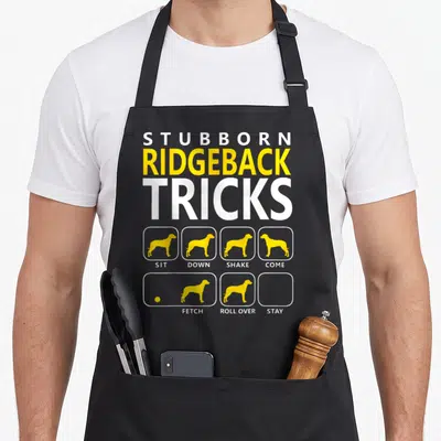 Vintage rhodesian ridgeback apron - Stubborn Ridgeback Tricks text with yellow dog silhouettes and sit down shake come — shown on a black cotton canvas apron against a white background