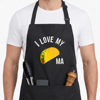 Taco truck apron - Cartoon taco with yellow shell and colorful fillings under arched white text reading I LOVE MY MA — shown on a black cotton canvas apron against a white background