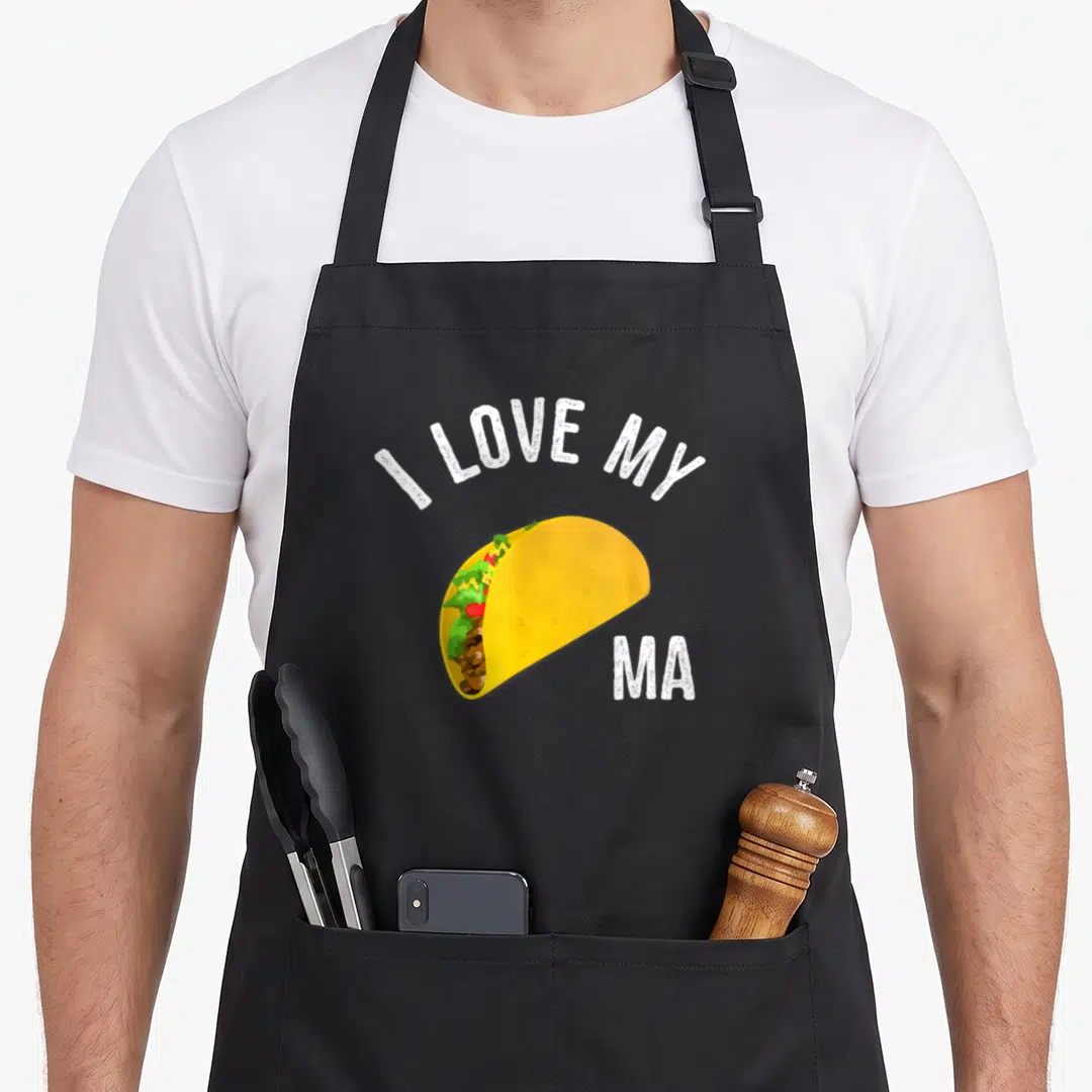 Taco truck apron - Cartoon taco with yellow shell and colorful fillings under arched white text reading I LOVE MY MA — shown on a black cotton canvas apron against a white background