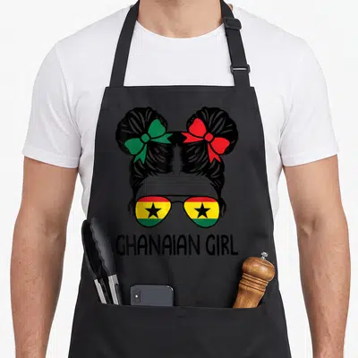 Ghanaian messy aprons - Cartoon girl head with buns, flag sunglasses, and text GHANAIAN GIRL — shown on a black cotton canvas apron against a white background
