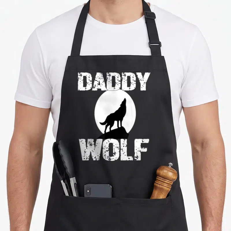 Wolf grill daddy apron - DADDY WOLF text framing a white moon with a black wolf silhouette on a rock — shown on a black cotton canvas apron against a white background