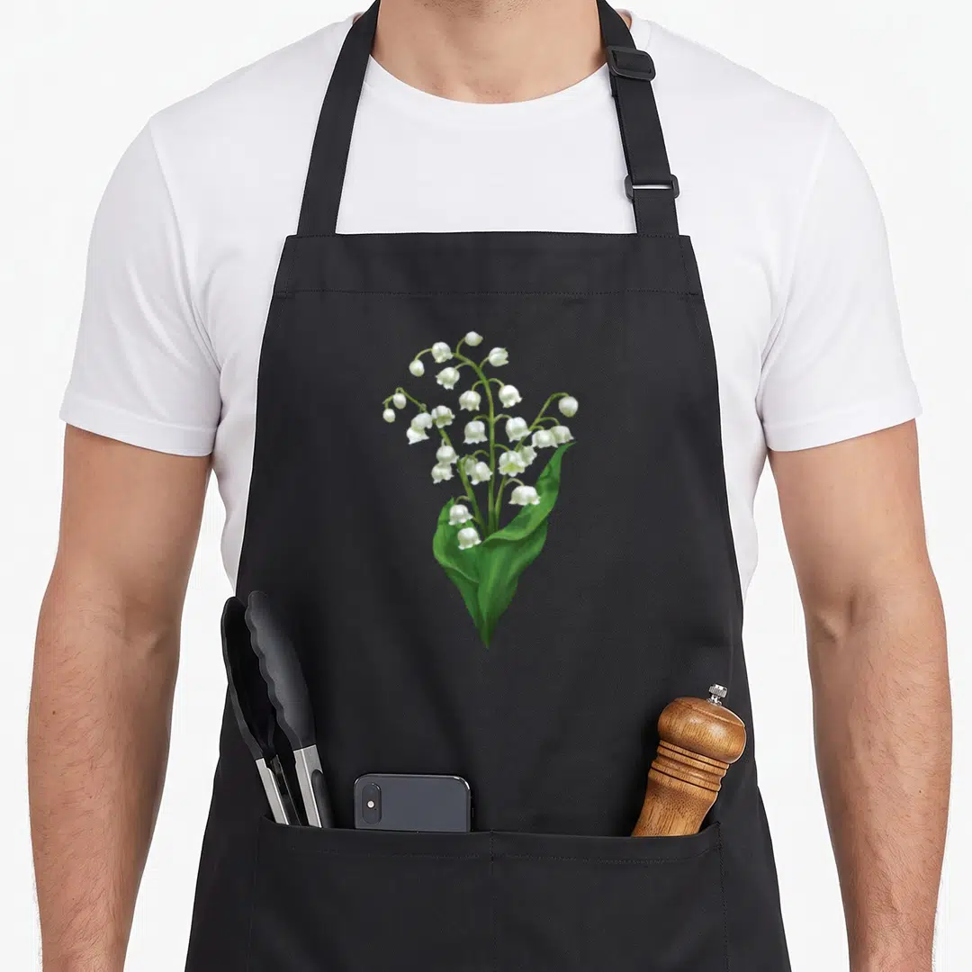 Spring white lily apron - Realistic lily of the valley stem with white bells and large green leaves on black fabric — shown on a black cotton canvas apron against a white background