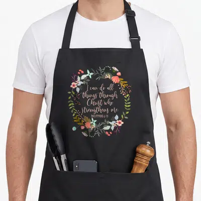 Philippian floral apron - Floral wreath surrounding pinkish-tan script I can do all things through Christ who — shown on a black cotton canvas apron against a white background