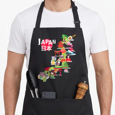 Travel pride apron - Green Japan map with JAPAN text, lucky cat, sushi, pagoda, and cherry blossom icons in cartoon — shown on a black cotton canvas apron against a white background