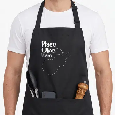 Music accessory ukulele apron - White Place Uke Here text with arrow pointing to dashed ukulele outline — shown on a black cotton canvas apron against a white background