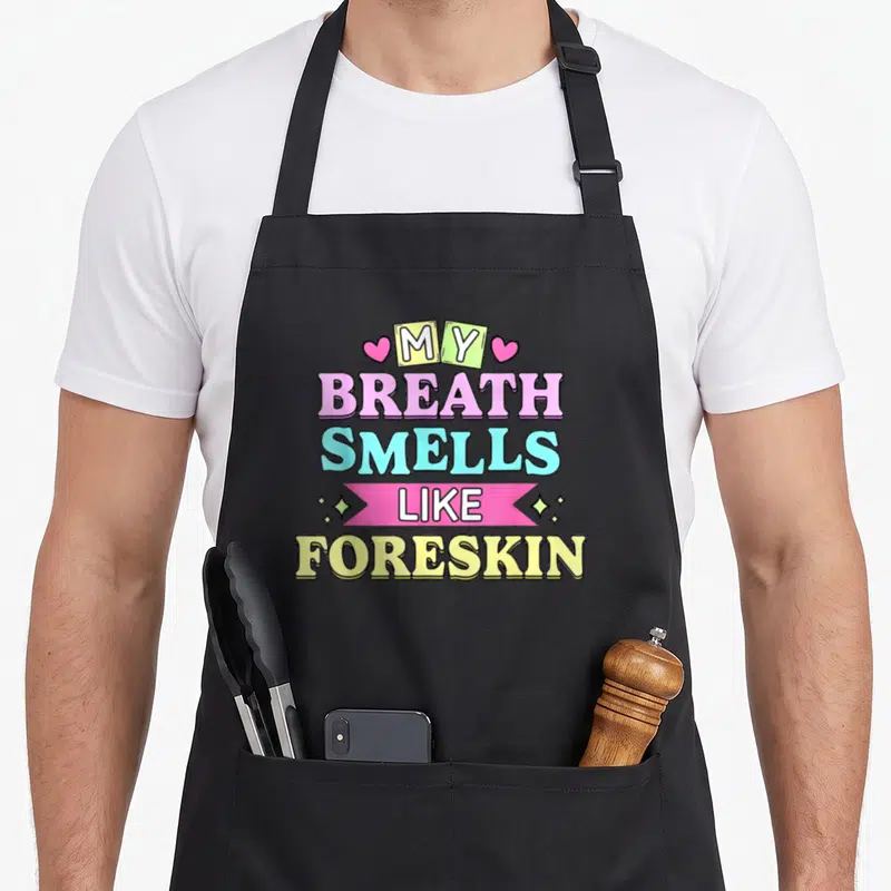 Breath the dirty apron - Stacked text My Breath Smells Like Foreskin with hearts and pink ribbon in pink yellow blue — shown on a black cotton canvas apron against a white background