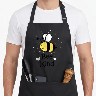 Bumble bee apron - Cartoon bee with Bee Kind text and two white hearts surrounded by dots — shown on a black cotton canvas apron against a white background