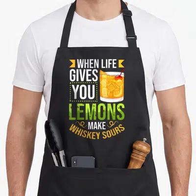 Whiskey recipe apron - Text when life gives you lemons make whiskey sours next to cocktail glass with ice and cherry — shown on a black cotton canvas apron against a white background