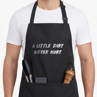 Dirt tough mudder apron - White distressed text A Little Dirt Never Hurt on black fabric — shown on a black cotton canvas apron against a white background