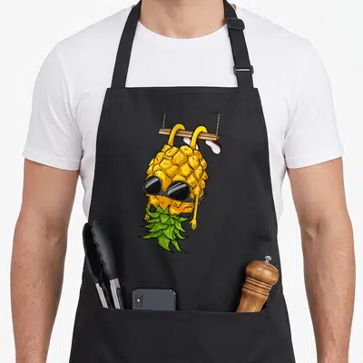 Cartoon pineapple apron - Upside down yellow pineapple wearing sunglasses holding green leaves hanging from wooden plank — shown on a black cotton canvas apron against a white background