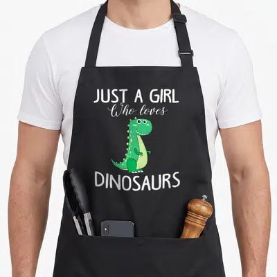 Cute dinosaur t-rex apron - Green cartoon dinosaur between white text JUST A GIRL Who loves DINOSAURS — shown on a black cotton canvas apron against a white background