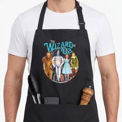 Funny wizard off see apron - Vintage Wizard of Oz group photo in circle with blue title text and yellow brick road path — shown on a black cotton canvas apron against a white background