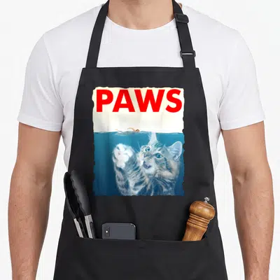 Parody shark cat apron - Red PAWS text above a photo of a swimmer and a cat reaching up from blue water — shown on a black cotton canvas apron against a white background