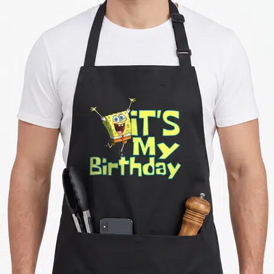 Funny squarepant birthday apron - SpongeBob with raised arms next to stacked IT'S My Birthday text in yellow and blue — shown on a black cotton canvas apron against a white background