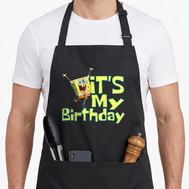 Funny squarepant birthday apron - SpongeBob with raised arms next to stacked IT'S My Birthday text in yellow and blue — shown on a black cotton canvas apron against a white background