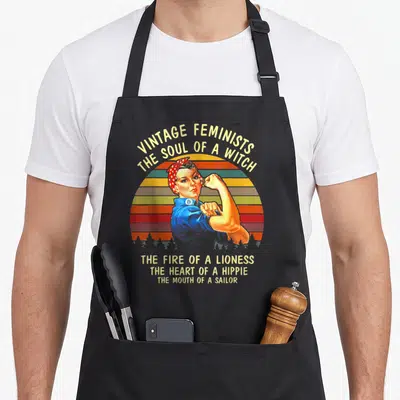 Feminist witch apron - Retro woman flexing arm under arched Vintage Feminists text and stacked quotes over striped sun — shown on a black cotton canvas apron against a white background