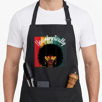 Apologetic african american aprons - Cartoon woman with afro and heart sunglasses reading Unapologetically DOPE — shown on a black cotton canvas apron against a white background