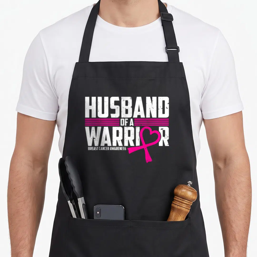 Husband breast cancer awareness aprons - Distressed HUSBAND OF A WARRIOR text with pink ribbon replacing the O and five — shown on a black cotton canvas apron against a white background