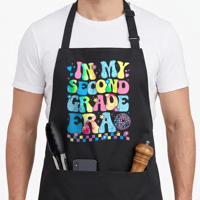 Era groovy teacher apron - Retro text IN MY SECOND GRADE ERA with disco ball, flowers, hearts, and checkerboard strip — shown on a black cotton canvas apron against a white background
