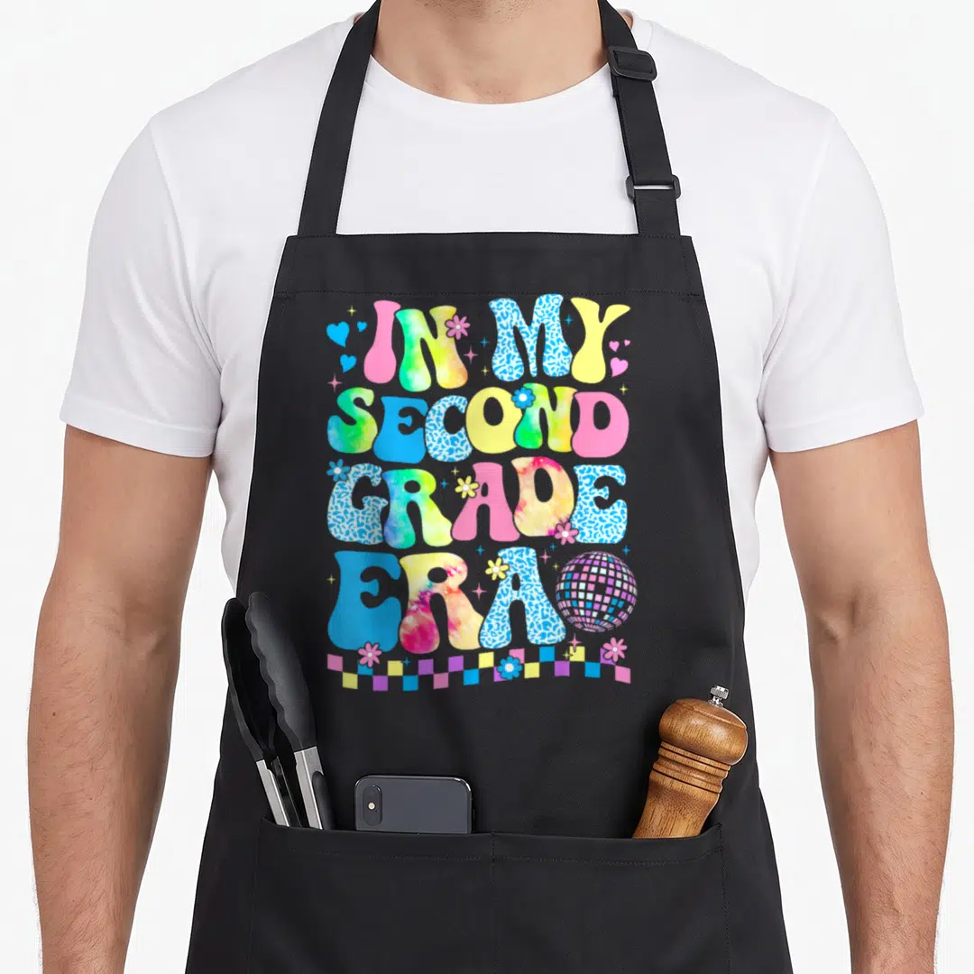 Era groovy teacher apron - Retro text IN MY SECOND GRADE ERA with disco ball, flowers, hearts, and checkerboard strip — shown on a black cotton canvas apron against a white background