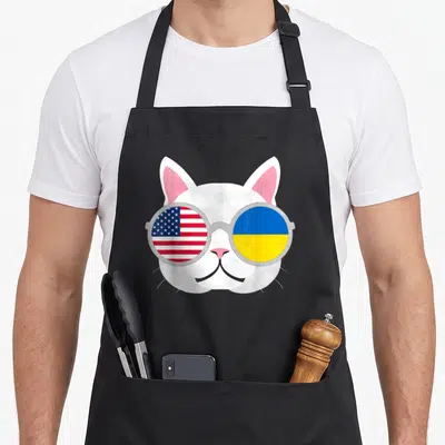 Proud ukrainian american flag apron - White cartoon cat head wearing grey round sunglasses with USA and Ukraine flag — shown on a black cotton canvas apron against a white background