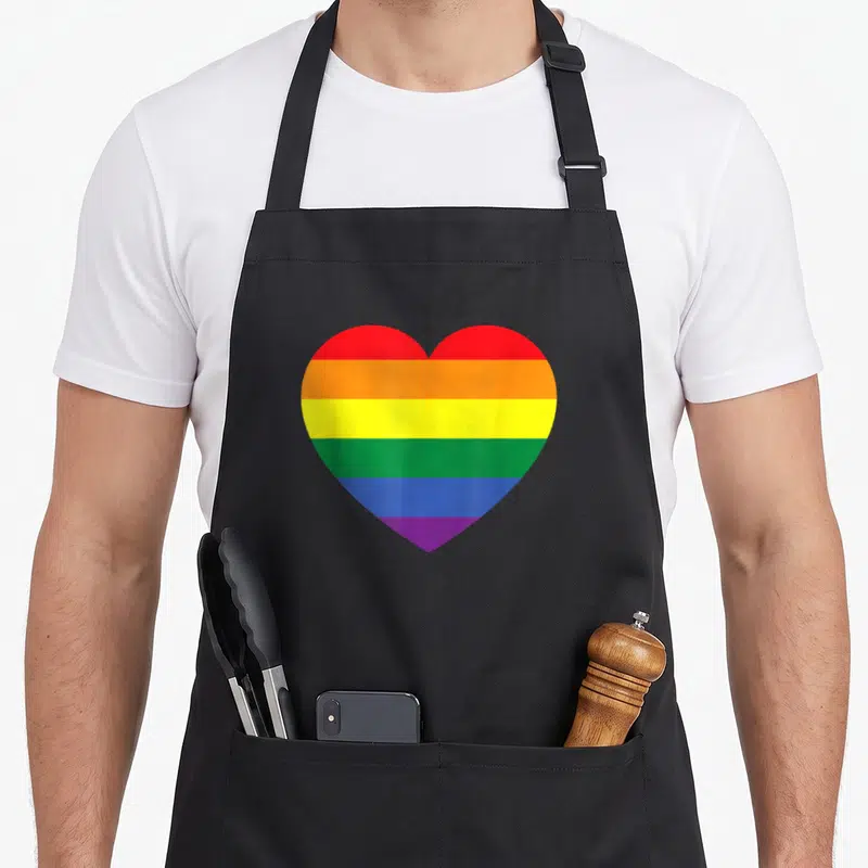 Colored gay pride apron - Rainbow heart with red orange yellow green blue purple stripes on black background — shown on a black cotton canvas apron against a white background