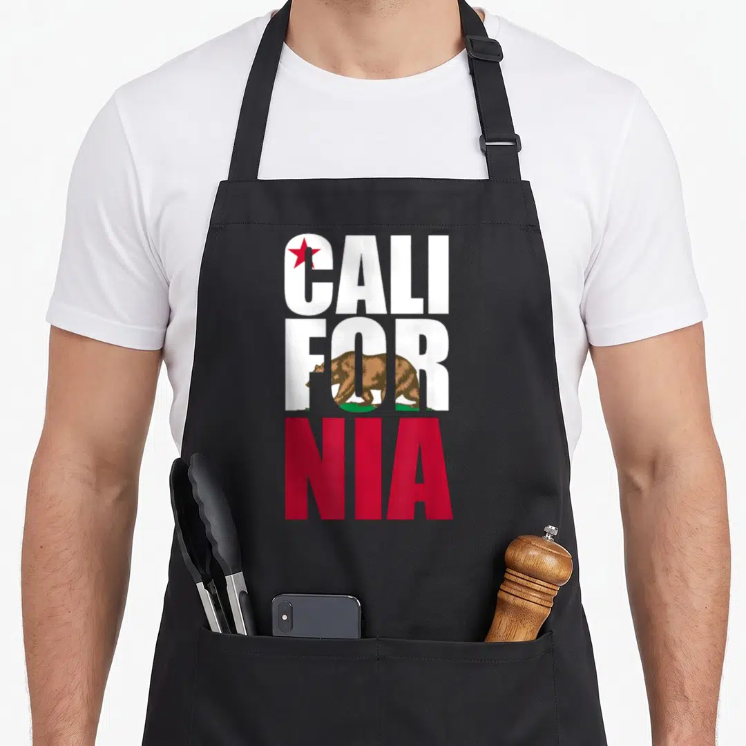 Cute california bear apron - Split CALIFORNIA text with brown bear on green grass and red star in white C above red NIA — shown on a black cotton canvas apron against a white background