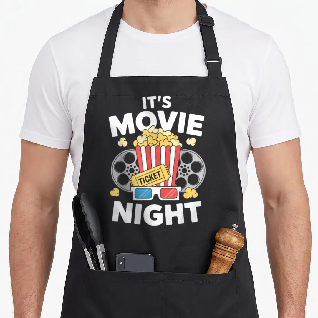Watch family aprons - Cartoon popcorn bucket with film reels, ticket, and 3D glasses under IT'S MOVIE NIGHT text — shown on a black cotton canvas apron against a white background