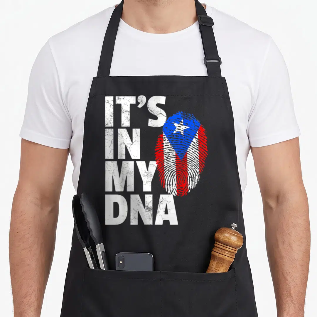 Puerto dna apron - Stacked distressed text IT'S IN MY DNA beside a red white blue fingerprint shaped like a flag — shown on a black cotton canvas apron against a white background
