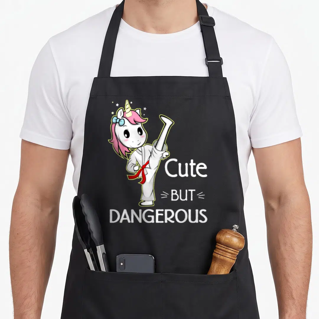 Karate unicorn apron - Cartoon unicorn in white gi with red belt kicking next to stacked text Cute But Dangerous — shown on a black cotton canvas apron against a white background
