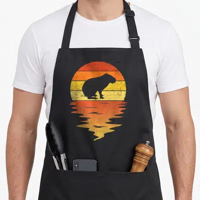 Capybara 70s apron - Grey capybara silhouette against red orange yellow striped sun with mirrored reflection — shown on a black cotton canvas apron against a white background