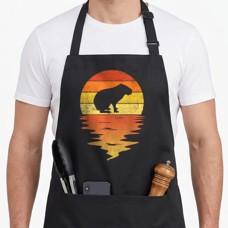 Capybara 70s apron - Grey capybara silhouette against red orange yellow striped sun with mirrored reflection — shown on a black cotton canvas apron against a white background