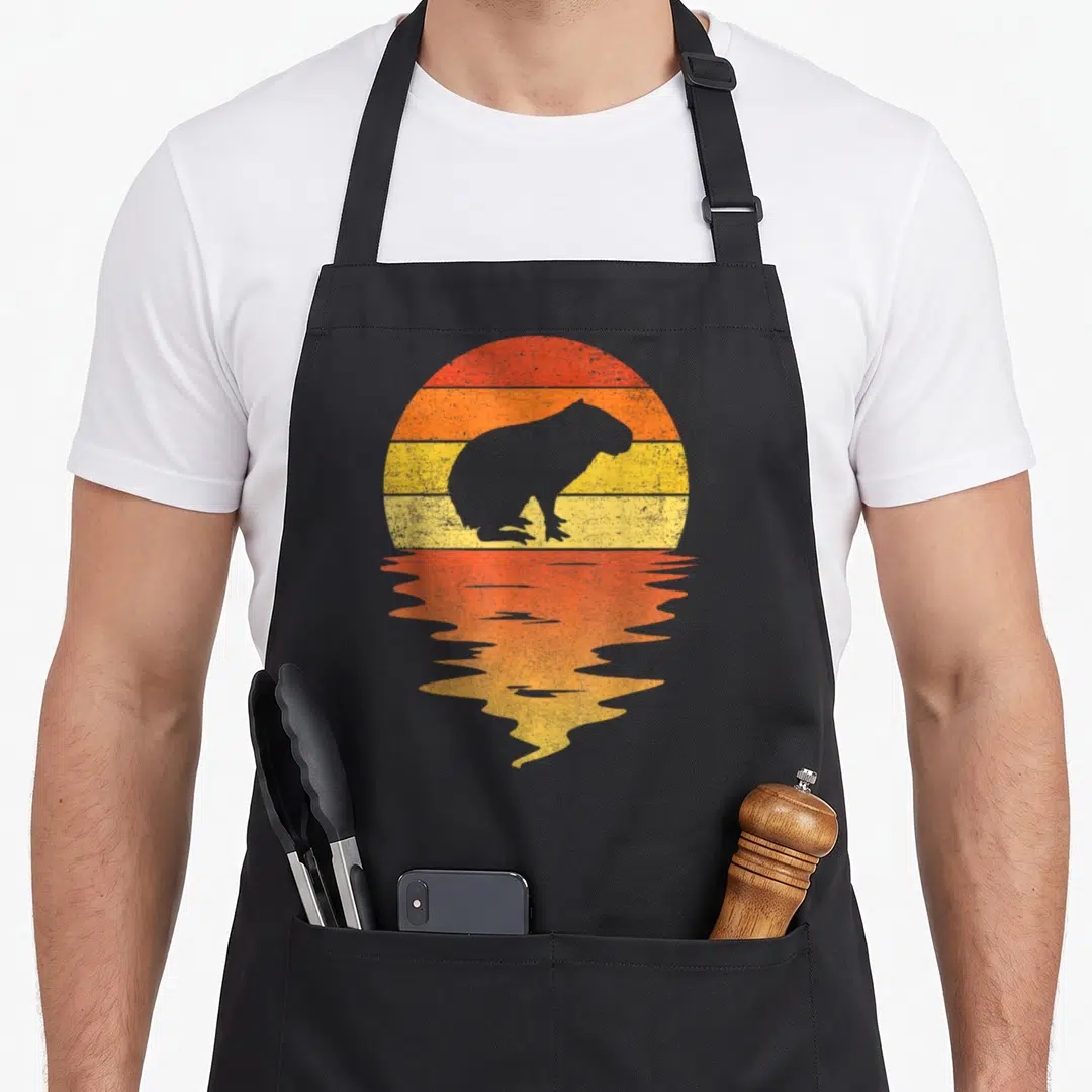 Capybara 70s apron - Grey capybara silhouette against red orange yellow striped sun with mirrored reflection — shown on a black cotton canvas apron against a white background