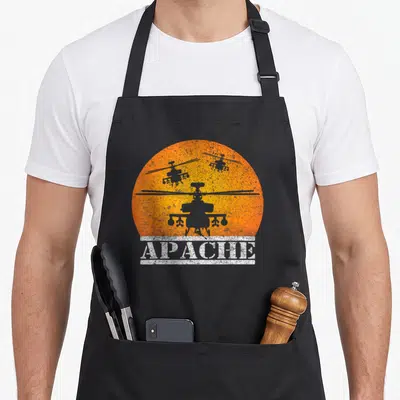 Apache flying apron - Three grey helicopter silhouettes against an orange sun with white APACHE text banner — shown on a black cotton canvas apron against a white background