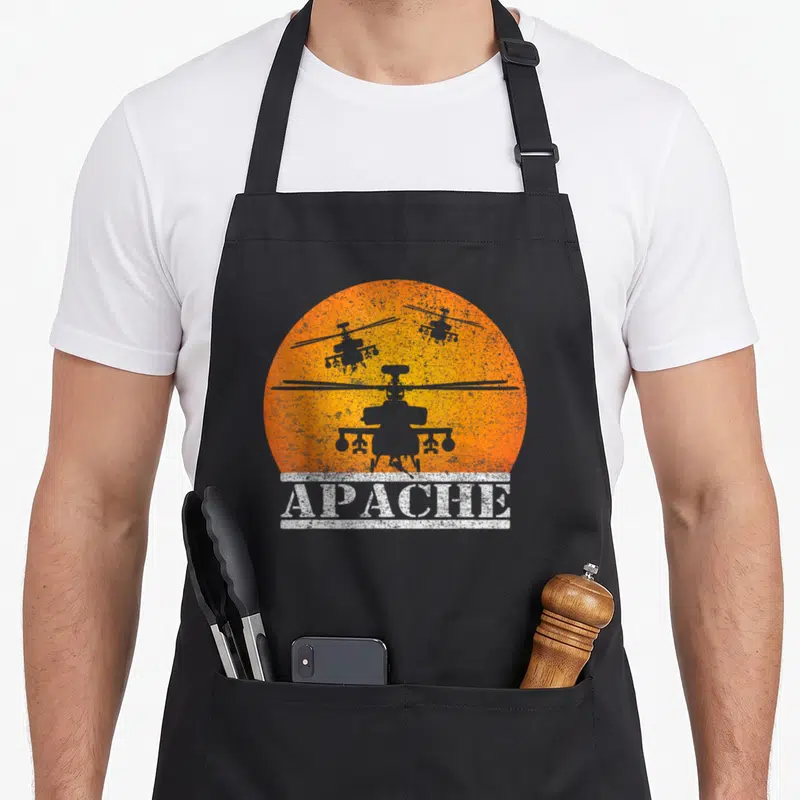 Apache flying apron - Three grey helicopter silhouettes against an orange sun with white APACHE text banner — shown on a black cotton canvas apron against a white background