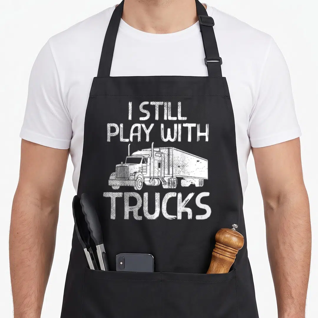 Play trucking semi truck apron - White distressed text reading I STILL PLAY WITH TRUCKS around a line drawing — shown on a black cotton canvas apron against a white background