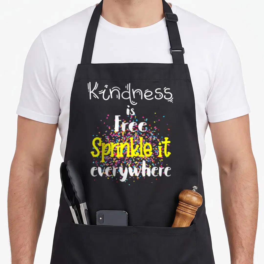 Kind sprinkle apron - Stacked text reading kindness is free sprinkled with colorful dots on black bib fabric — shown on a black cotton canvas apron against a white background