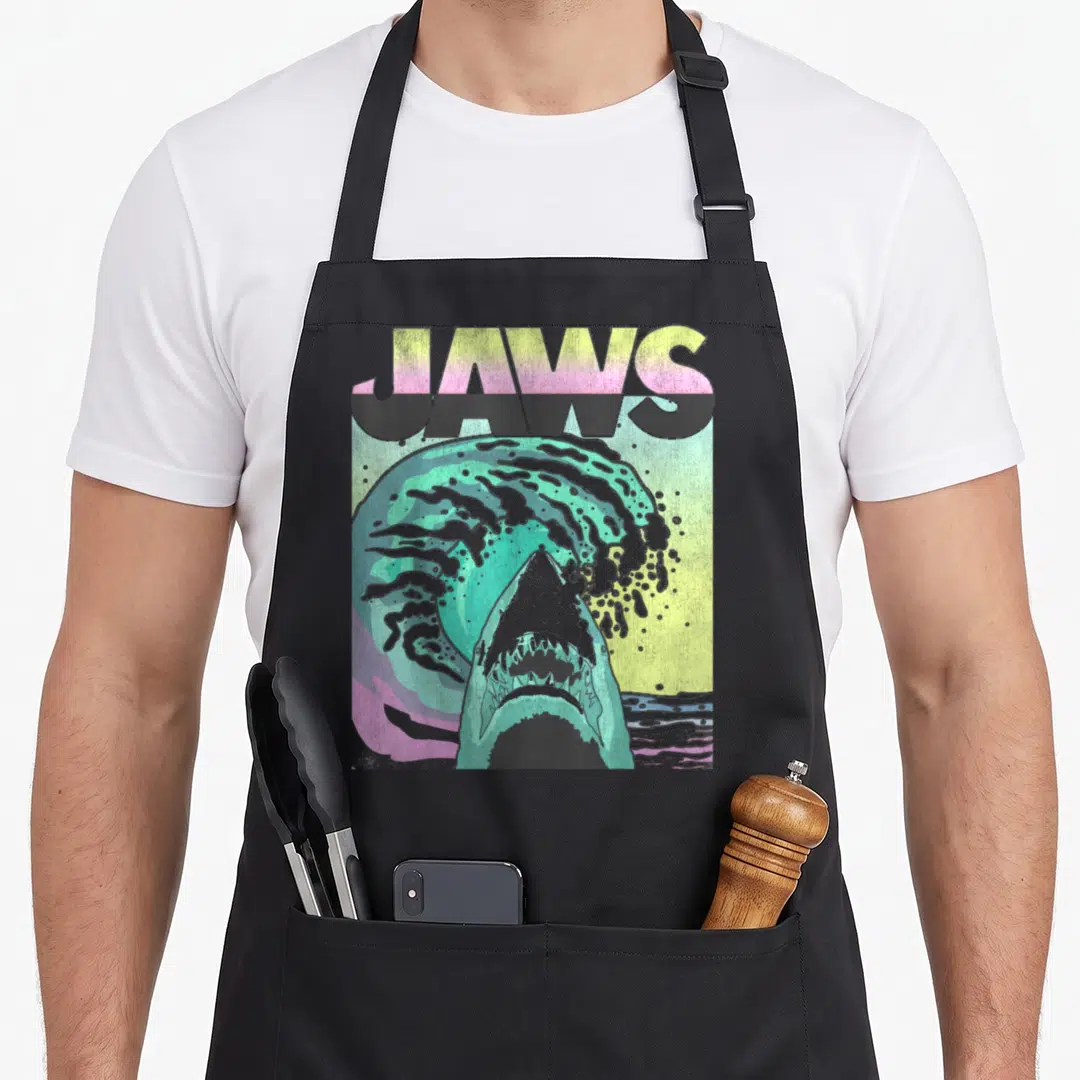 Poster wave apron - JAWS text above shark emerging from teal wave in retro pink yellow grey — shown on a black cotton canvas apron against a white background