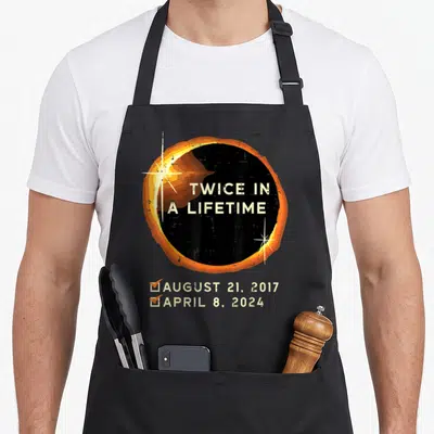 Lifetime april apron - Orange celestial circle with TWICE IN A LIFETIME text and checked dates for 2017 and 2024 — shown on a black cotton canvas apron against a white background