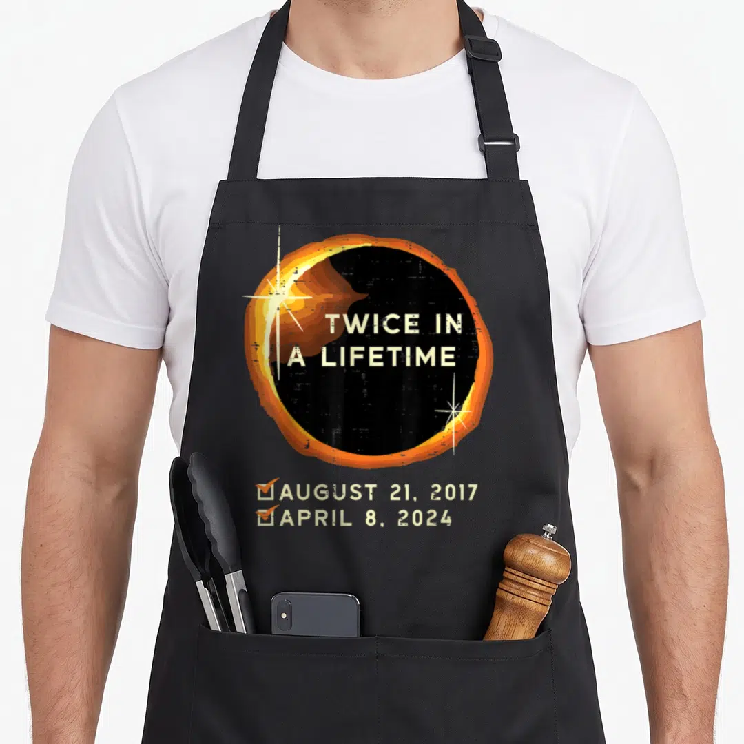 Lifetime april apron - Orange celestial circle with TWICE IN A LIFETIME text and checked dates for 2017 and 2024 — shown on a black cotton canvas apron against a white background