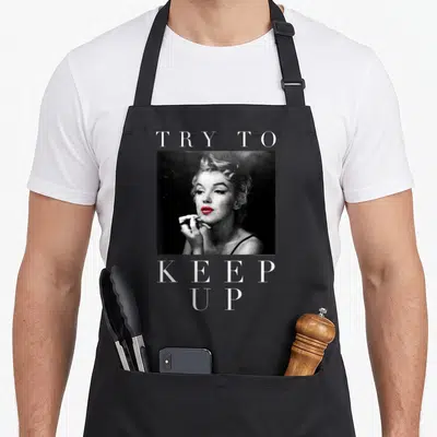 Marilyn sampling apron - TRY TO KEEP UP text framing grayscale woman photo with red lips — shown on a black cotton canvas apron against a white background
