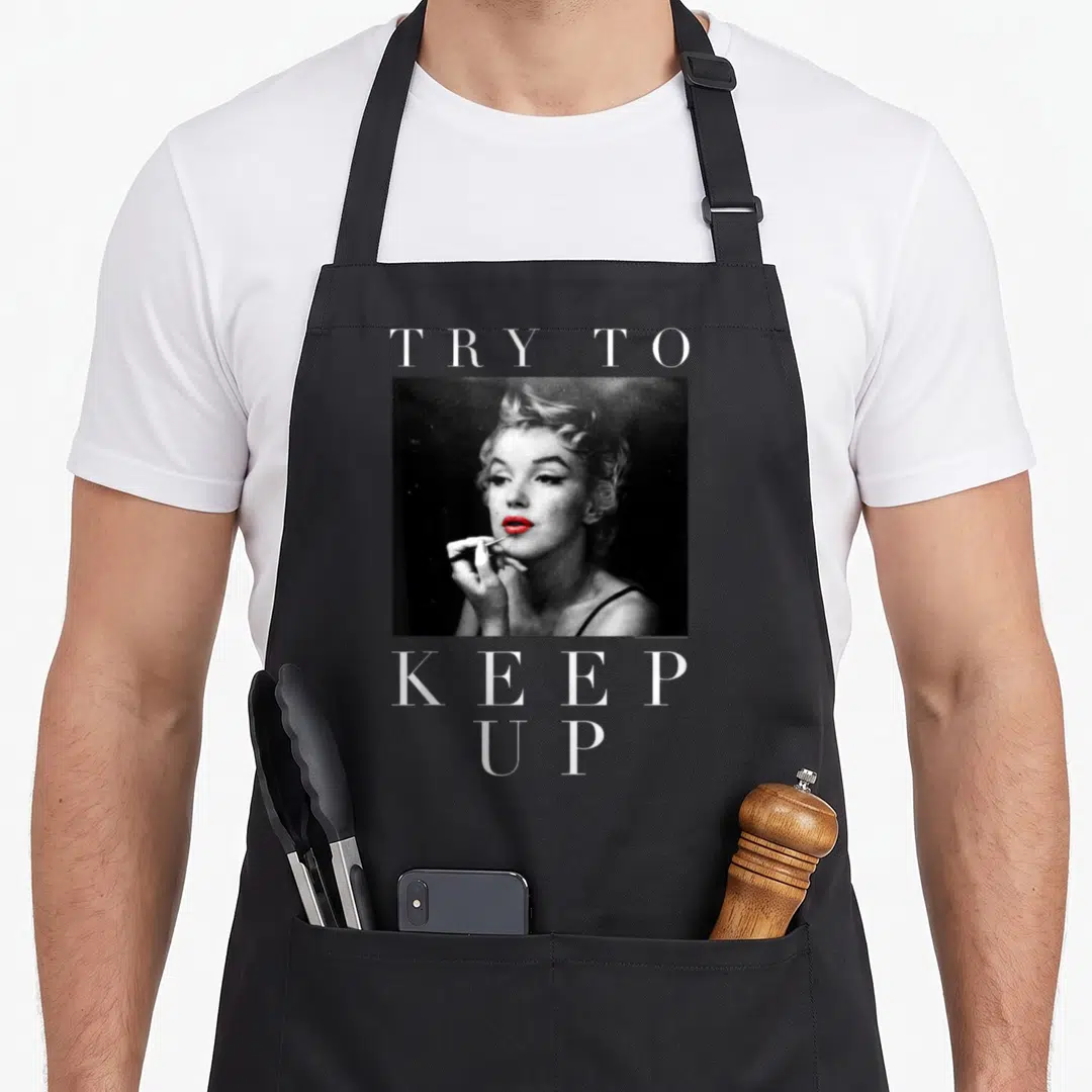Marilyn sampling apron - TRY TO KEEP UP text framing grayscale woman photo with red lips — shown on a black cotton canvas apron against a white background