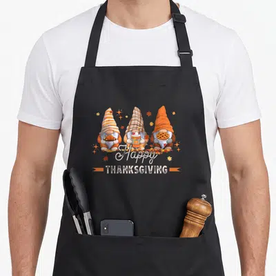 Gnome thanksgiving day aprons - Three gnomes holding turkey, drink, and pie below Happy THANKSGIVING banner in autumn — shown on a black cotton canvas apron against a white background
