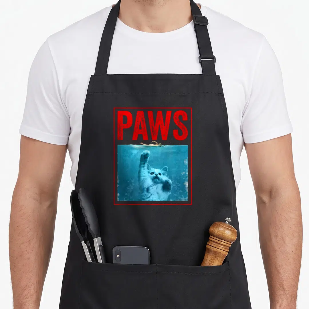 Paw kitten apron - Red PAWS text above white cat reaching up underwater below floating swimmer in blue water inside red — shown on a black cotton canvas apron against a white background