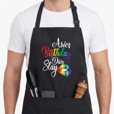 Mythical birthday apron - Stacked Aries Birthday Diva Slay text with rainbow lips graphic — shown on a black cotton canvas apron against a white background