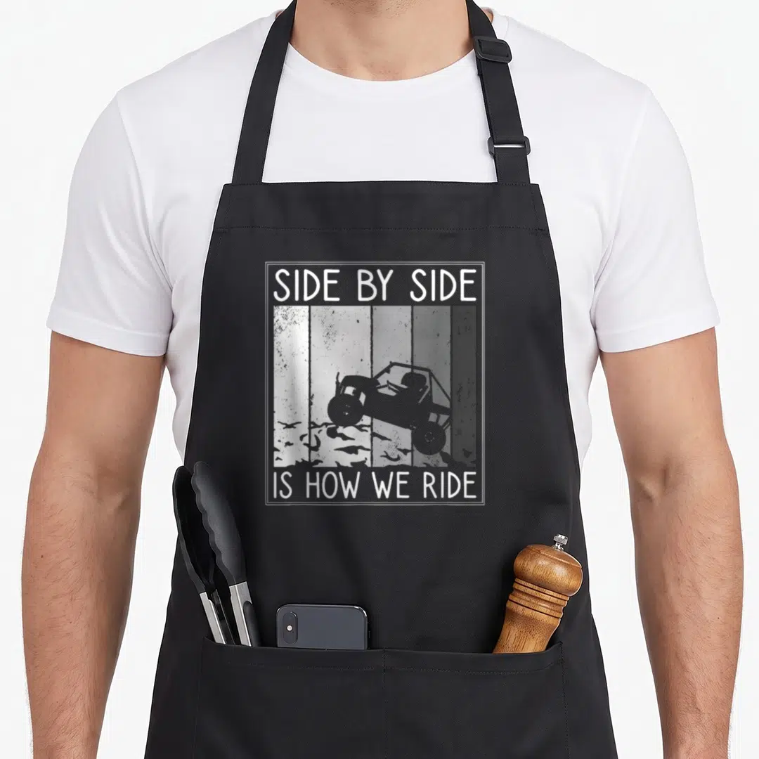 Ride by apron - Distressed UTV silhouette against grey vertical stripes with white text border reading Side By Side Is — shown on a black cotton canvas apron against a white background