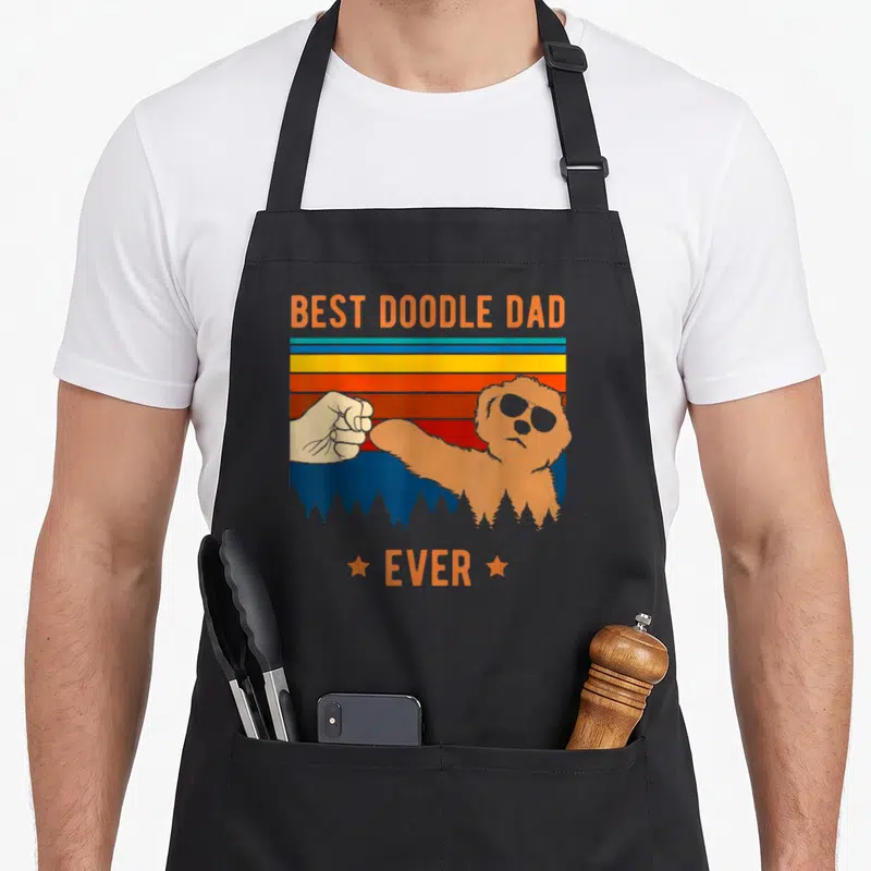 Goldendoodle labradoodle dog apron - Retro graphic of a tan dog with sunglasses fist-bumping a human hand against — shown on a black cotton canvas apron against a white background